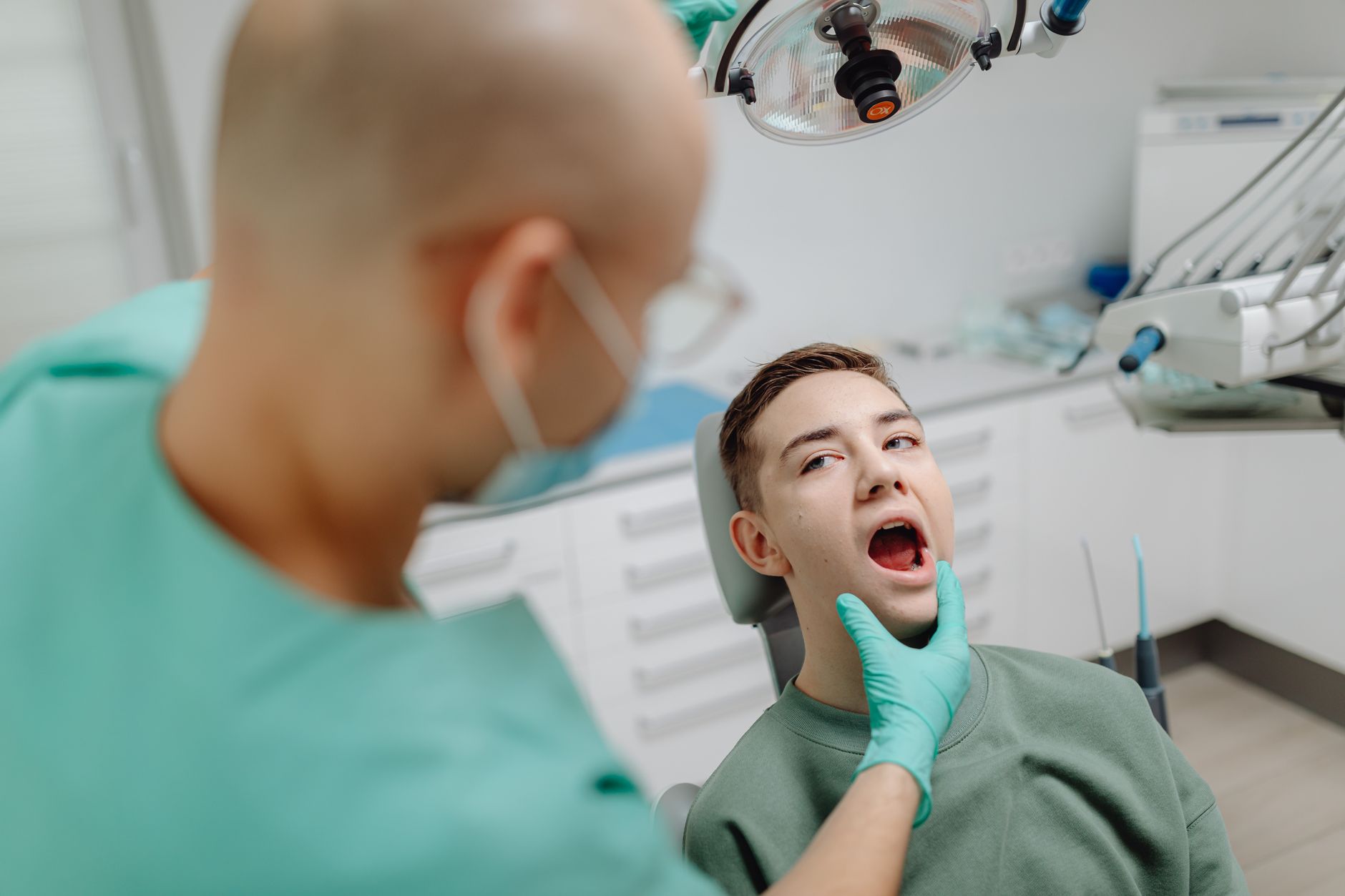 A dentist in mint green scrubs and a face mask leans over a teenage boy who is sitting back in a dental chair with his mouth open under the overhead light