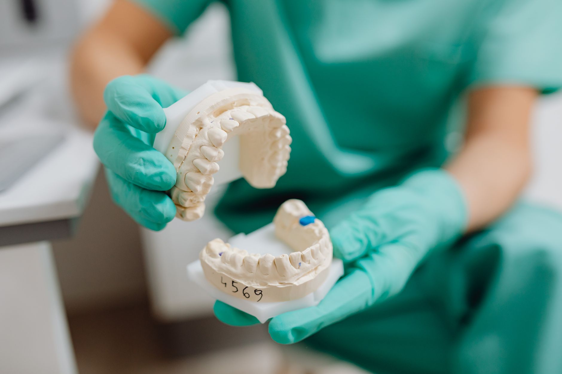 Gloved hands in green scrubs holding an upper and a lower plaster model of a set of teeth, the lower model marked with a handwritten case number