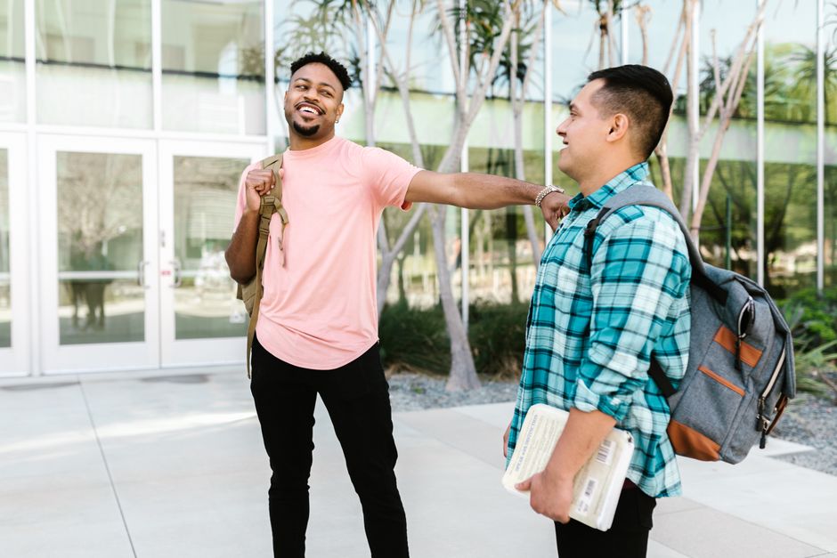 Two college students laughing together outside a glass-fronted campus building, both carrying backpacks and one holding a textbook