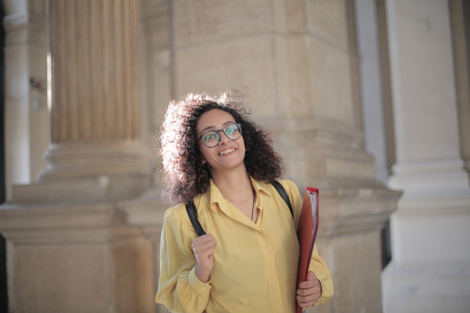 Smiling young woman with curly hair and glasses, wearing a yellow blouse and a backpack, holding a red folder in front of stone columns