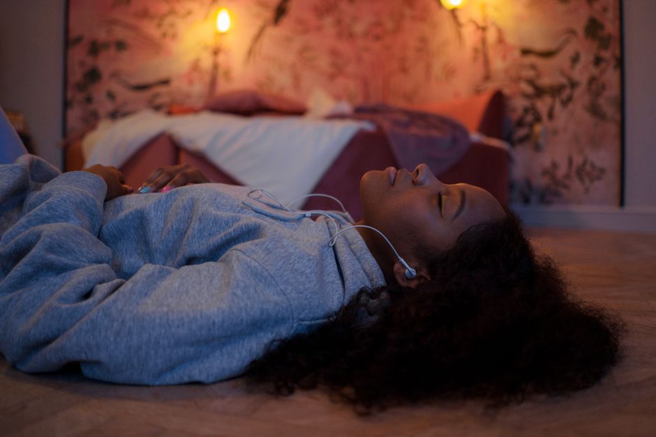 A woman with curly hair lying on the floor in a dim, warmly lit room with her eyes closed, wearing earbuds