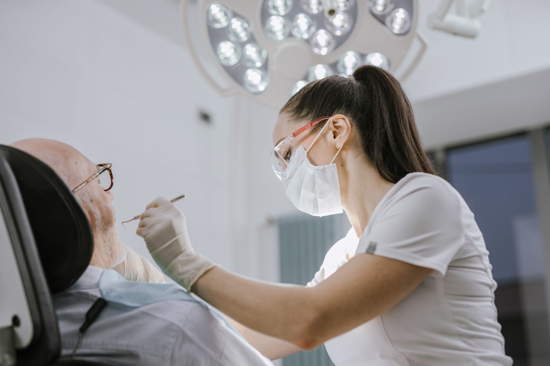 A dental professional in a mask and safety glasses examining an older patient reclined in a treatment chair under an overhead light