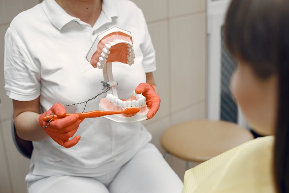 A dental professional in orange gloves holding a model of teeth and a toothbrush while a patient looks on