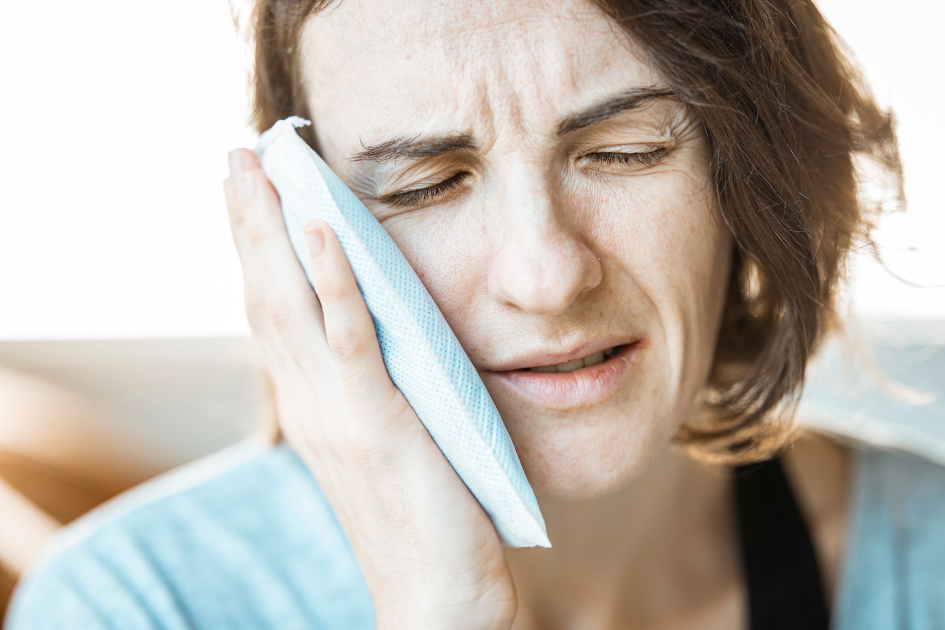 A woman with short brown hair winces with her eyes closed while holding a pale blue cold pack against her cheek and jaw.