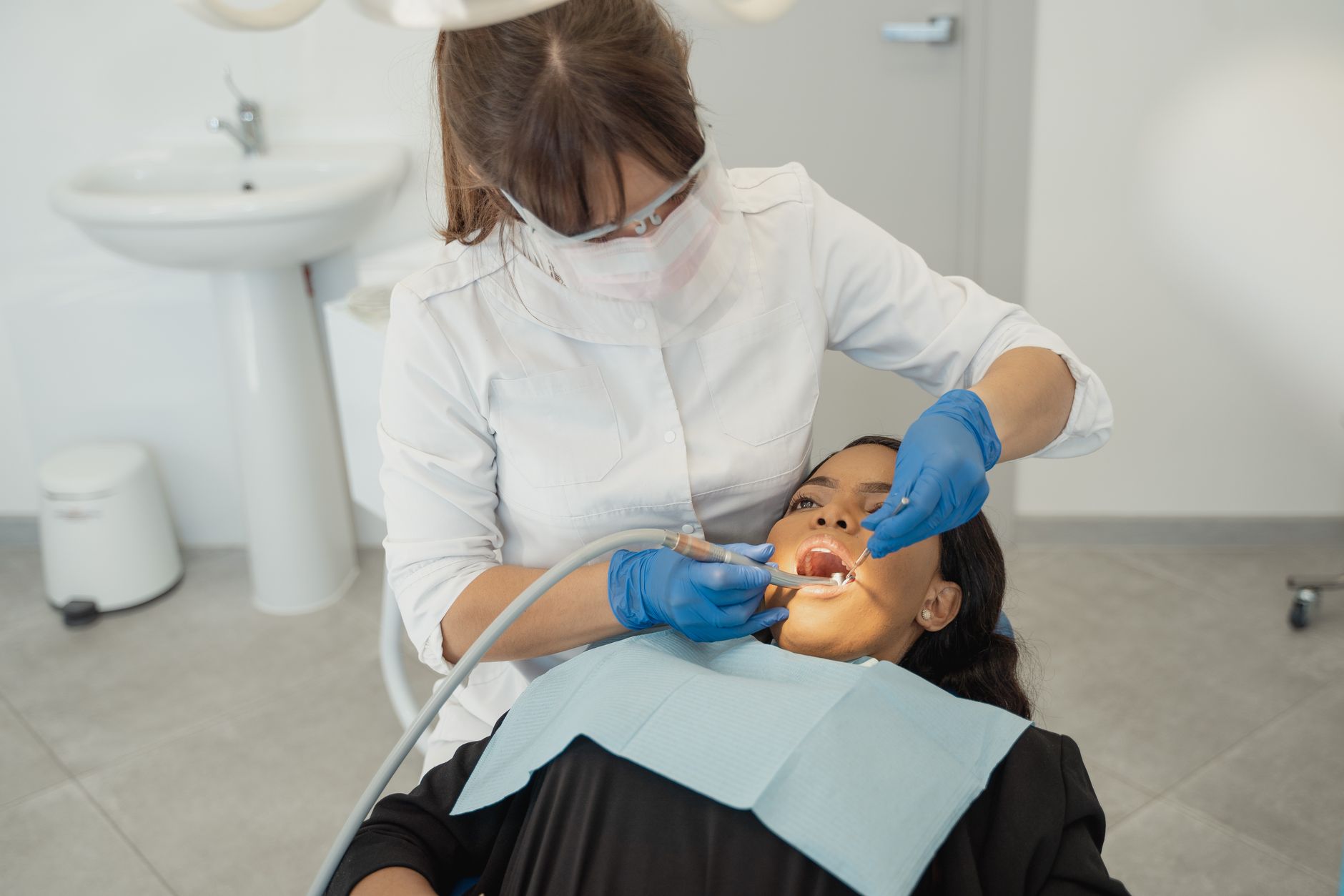 A dental clinician in a white coat, face shield and blue gloves examines the upper teeth of a patient reclined in the chair, using a mirror and a suction tip.
