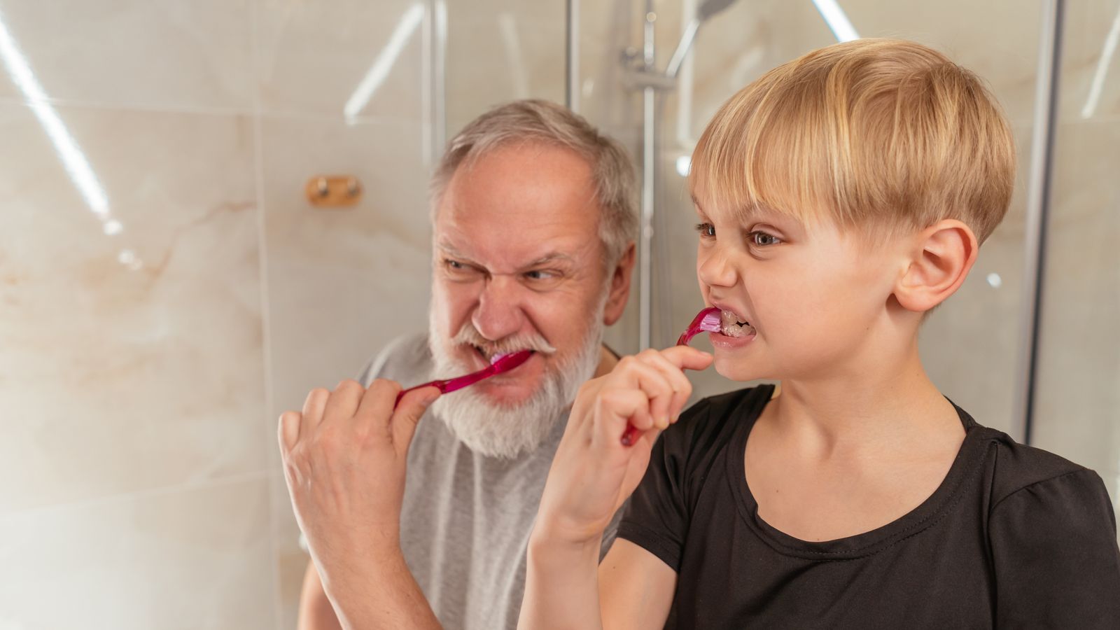 A grey-bearded older man and a blond boy pulling the same face while brushing their teeth side by side with pink toothbrushes in a bathroom