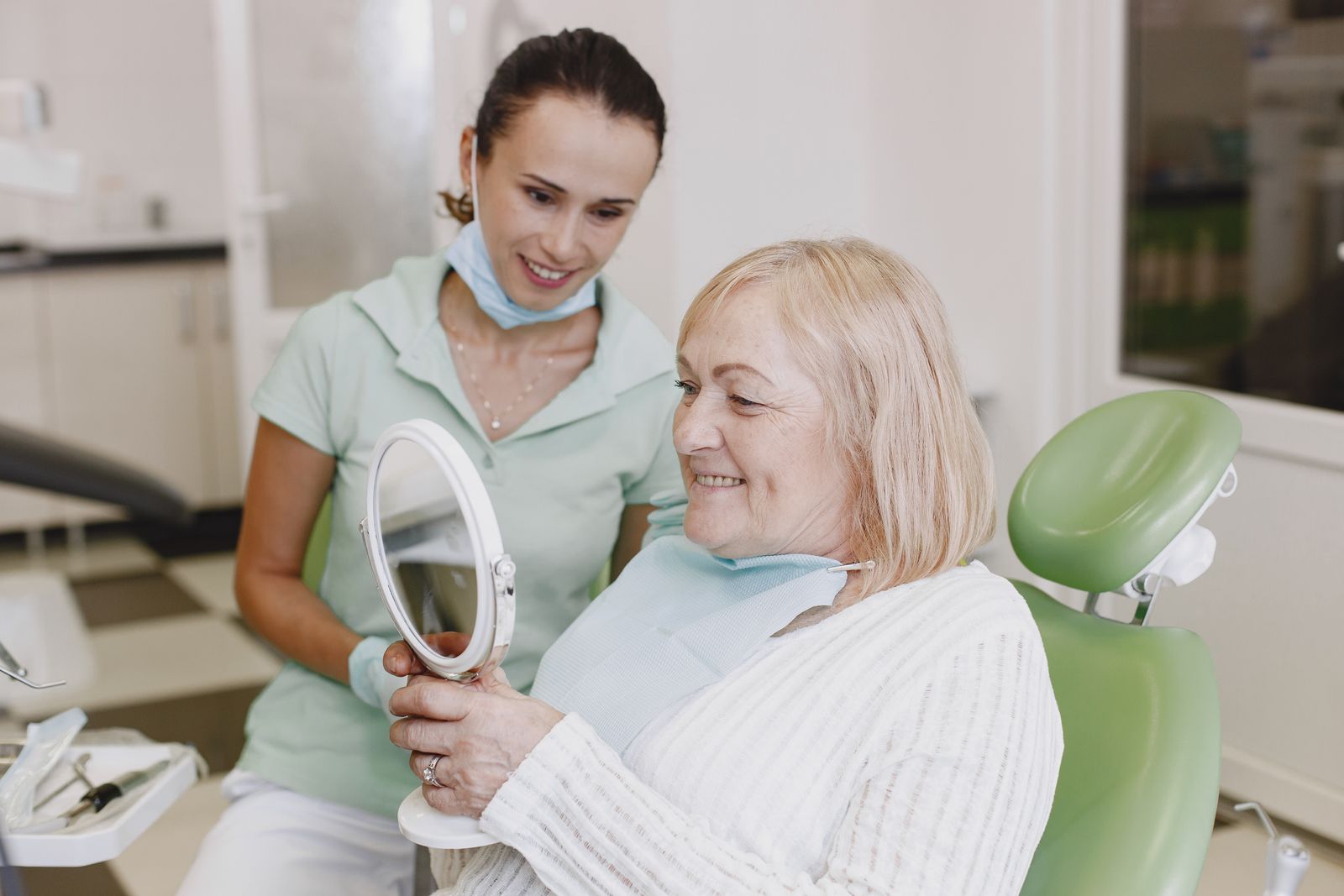 A smiling older woman in a green dental chair looking at her teeth in a hand mirror while a dental professional in mint scrubs smiles beside her