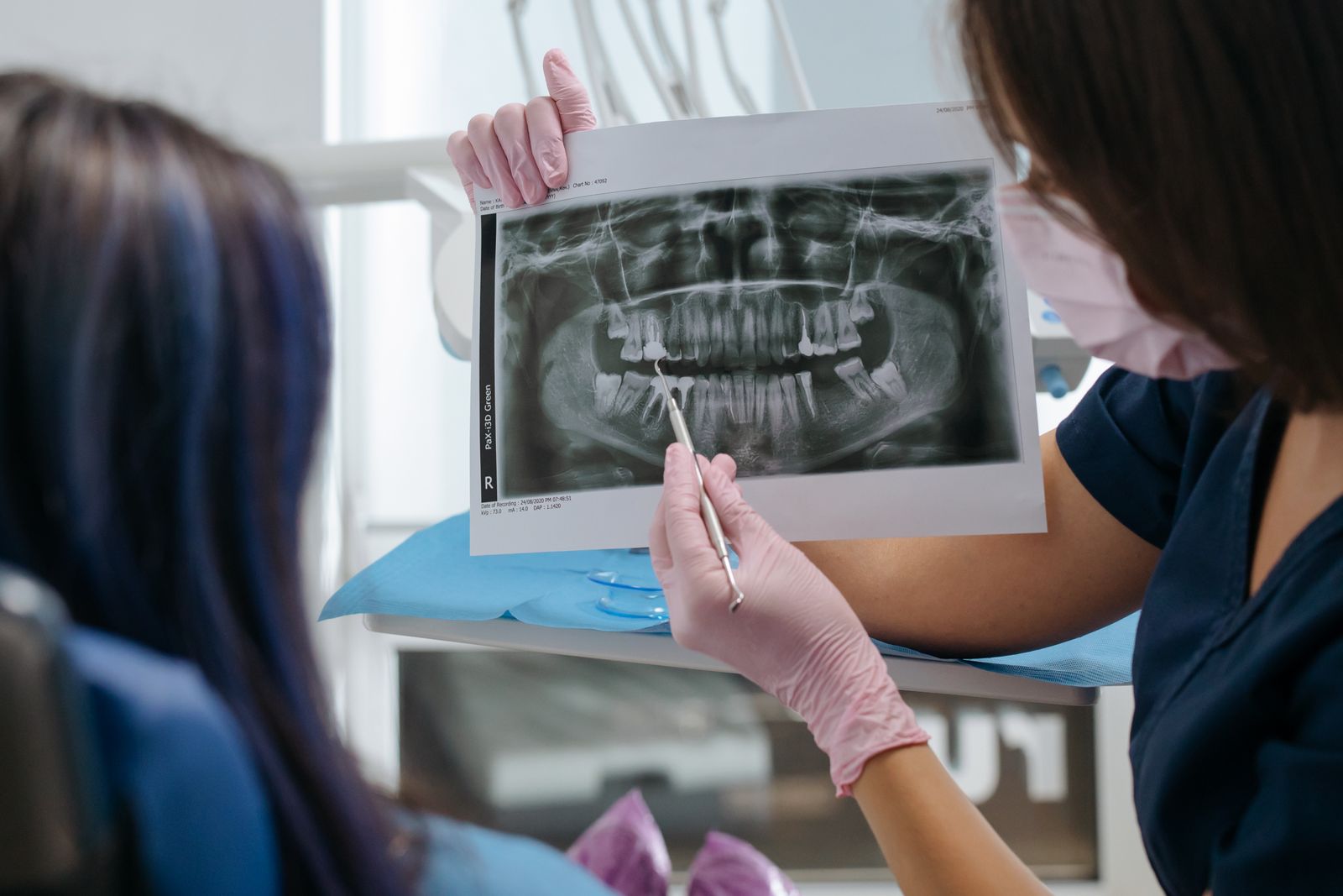 A dental professional in pink gloves and a mask pointing a dental probe at a panoramic X-ray held up for a seated patient